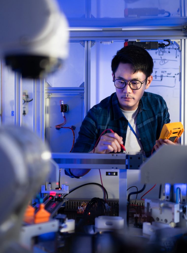 Young Asian male electrical engineer in glasses using a digital multimeter in hand checking voltage to fix an industrial machine with a blurred of automation robotic arm machine in the foreground.